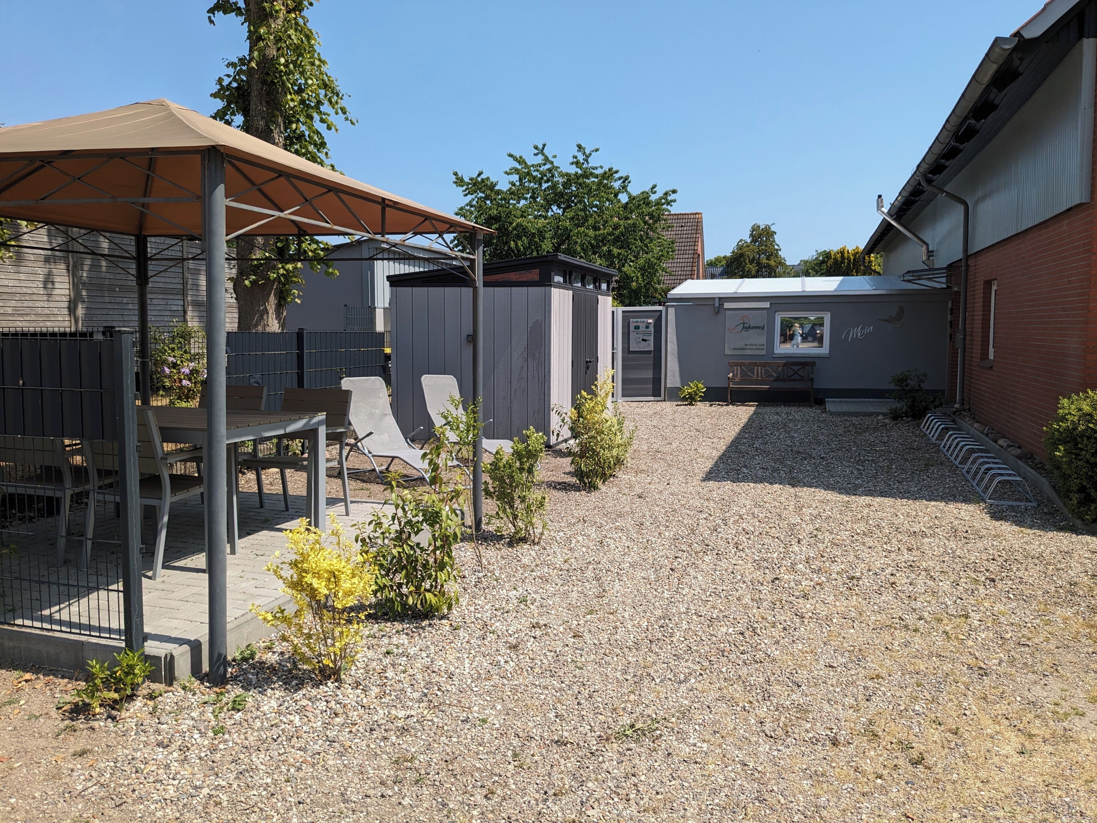 Ein überdachter Patio-Bereich mit Tisch und Stühlen, eine kleine Gartenhütte und ein gepflegter Schotterweg.