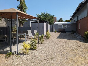 Ein überdachter Patio-Bereich mit Tisch und Stühlen, eine kleine Gartenhütte und ein gepflegter Schotterweg.
