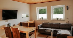 Ein Wohnzimmer mit einer Couch, einem Esstisch mit Stühlen, einem an der Wand befestigten Fernseher und zwei Fenstern mit Pflanzen.
