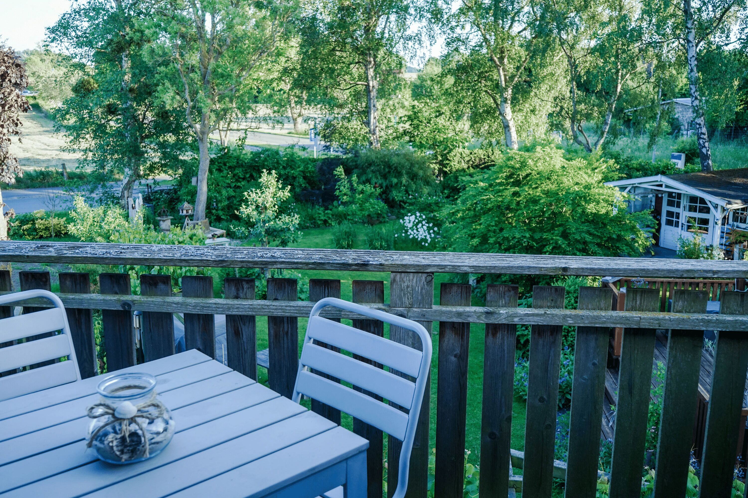 A wooden deck with a table and chairs, overlooking a lush garden and a small building.
