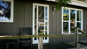 A wooden deck with a chair and a table, a house with a porch, and a large window reflecting trees. - Muskoka Cabin on Little Lake Port Severn (Port Severn)
