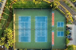 An aerial view of a tennis court complex with multiple courts, a parking area, and surrounding greenery.