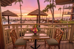 A balcony with a table and chairs, overlooking a pool and beach area with umbrellas and palm trees.
