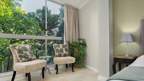 A room with a large window, a chair with floral upholstery, a small table, a potted plant, and a bedside lamp. - Sonwyk 9 (Cape Town)