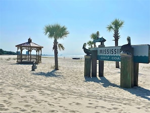 A beach with a wooden gazebo, palm trees, and a sign reading "MISSISSIPPI GULF COAST".