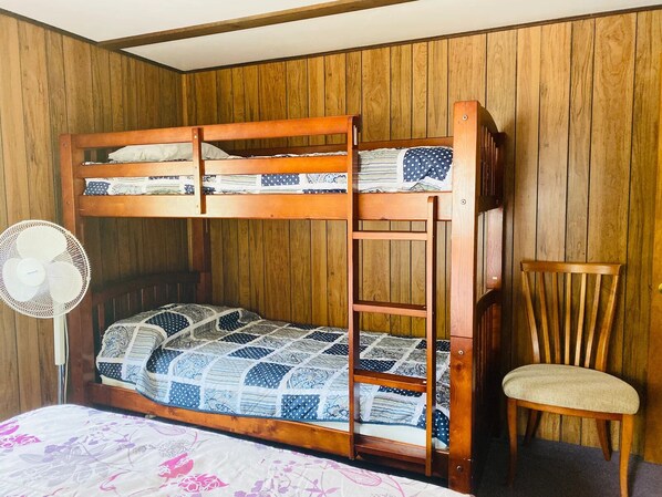 Un lit superposé avec une échelle, une chaise et un ventilateur sur pied, dans une pièce aux murs recouverts de panneaux de bois.