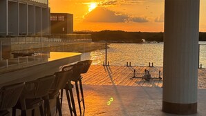 Un bar frente al agua con mesas al aire libre, vista del atardecer sobre el agua y un edificio moderno al fondo.