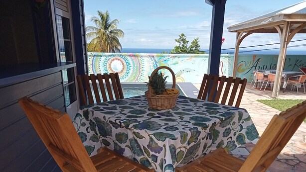 Una mesa de madera con mantel floral, rodeada de sillas de madera, ubicada en un patio con vista al océano y palmeras.