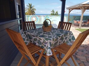 A wooden table with a floral tablecloth, surrounded by wooden chairs, set on a patio with a view of the ocean and palm trees. - AWMONY'KAZ: Charming bungalow with a pool and hydrotherapy jets. (Trois-Rivières)