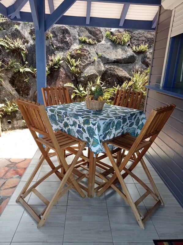 A covered patio with a table set for four, surrounded by wooden chairs, and a rocky backdrop with ferns. - AWMONY'KAZ: Charming bungalow with a pool and hydrotherapy jets. (Trois-Rivières)