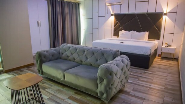 Un dormitorio moderno con un sofá gris capitonado, una mesita redonda pequeña, una cama con cabecera negra y una mesita de noche.