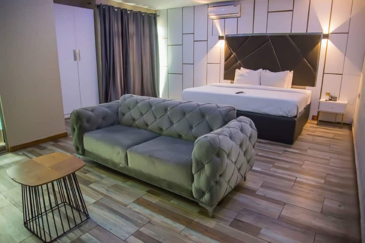 Un dormitorio moderno con un sofá gris capitonado, una mesita redonda pequeña, una cama con cabecera negra y una mesita de noche.