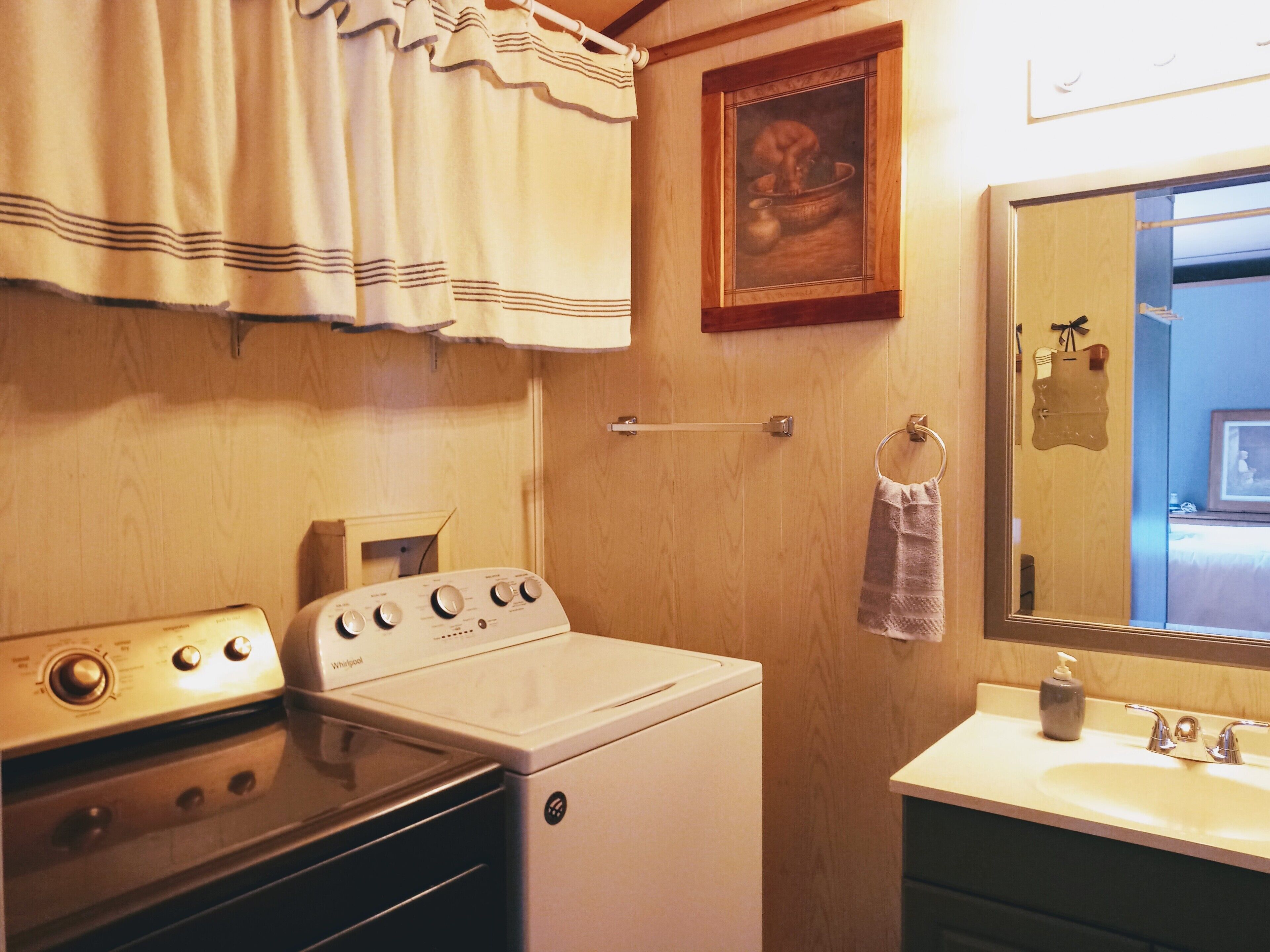 A laundry room with a Whirlpool washer and dryer, wooden cabinets, a sink with a mirror, and a framed painting on the wall.
