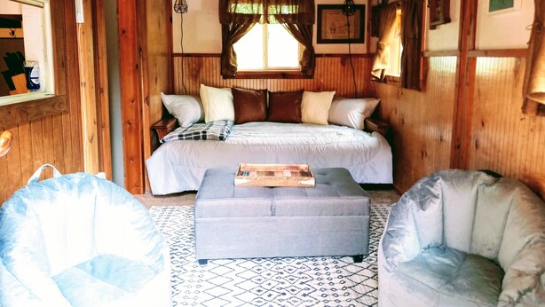 Une pièce chaleureuse aux murs en bois, avec un lit, un pouf gris et deux fauteuils.
