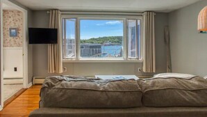 Quarto com cama, TV fixada na parede, janela com cortinas e vista para a água e edifícios.