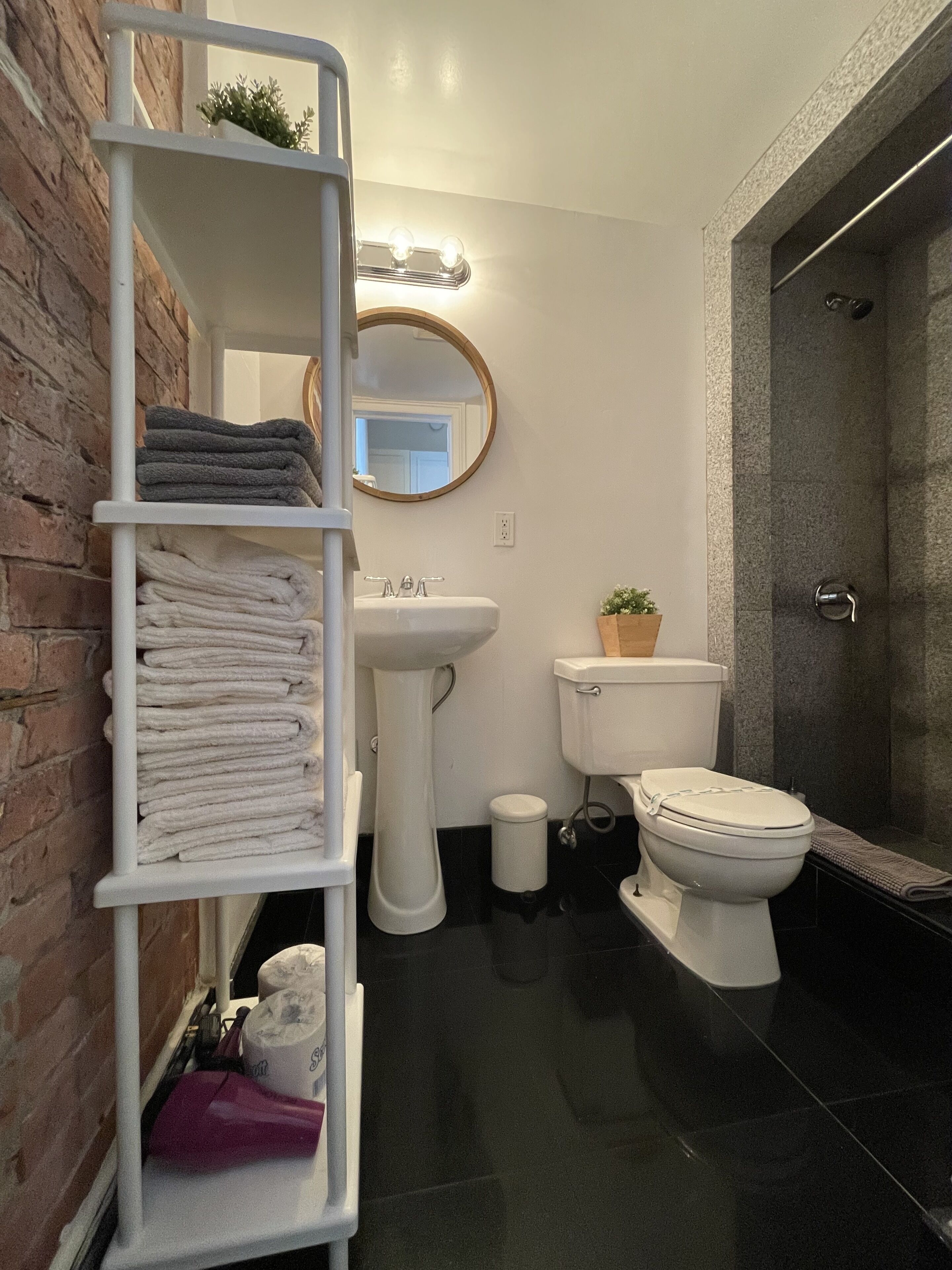 Un baño moderno con un lavamanos pedestal blanco, un inodoro y una zona de ducha. Hay repisas con toallas dobladas y una pequeña planta en maceta.