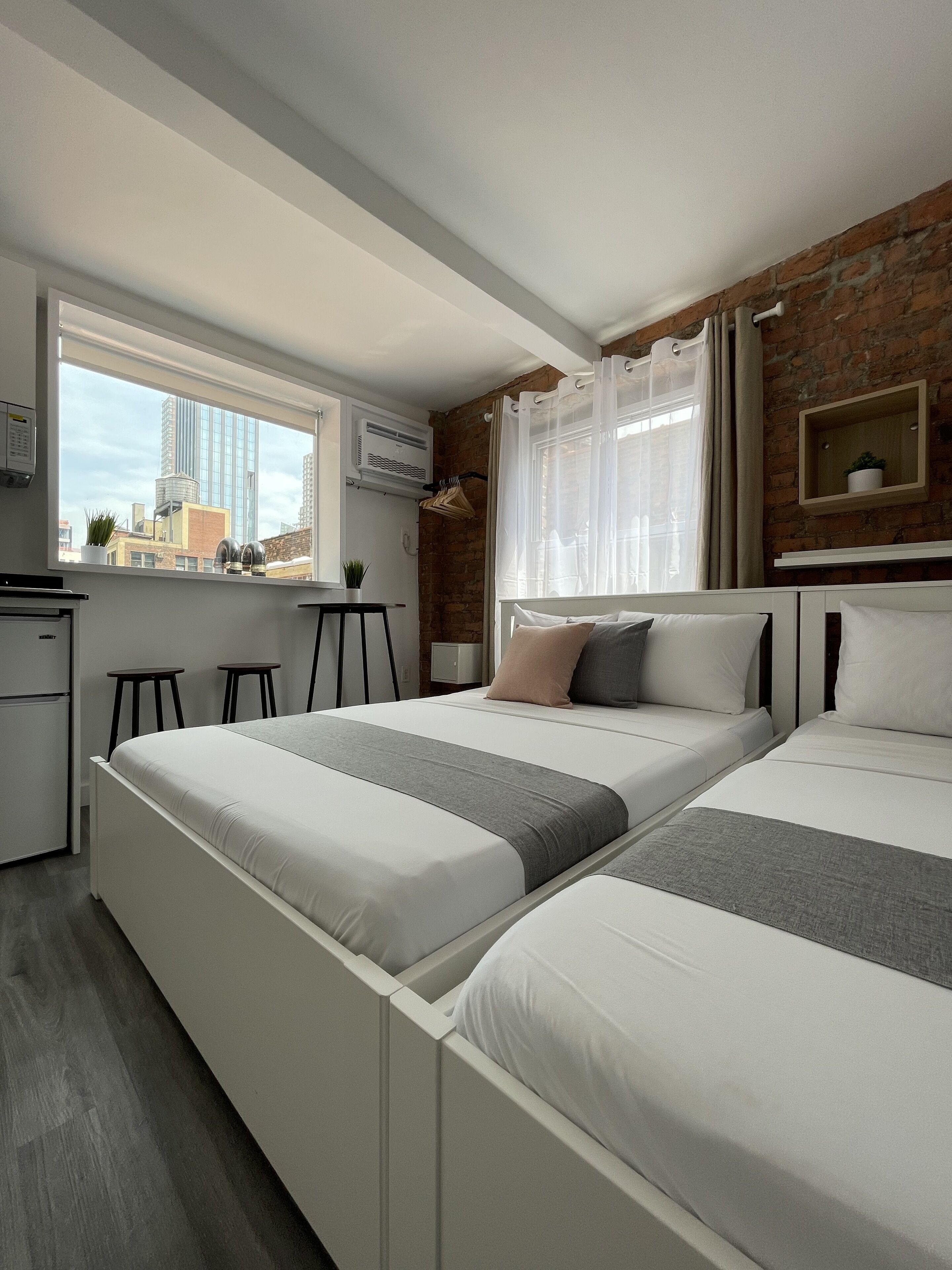 Een moderne slaapkamer met twee bedden, een bakstenen muur, een raam met gordijnen, een kleine plant en een witte inbouwkast.