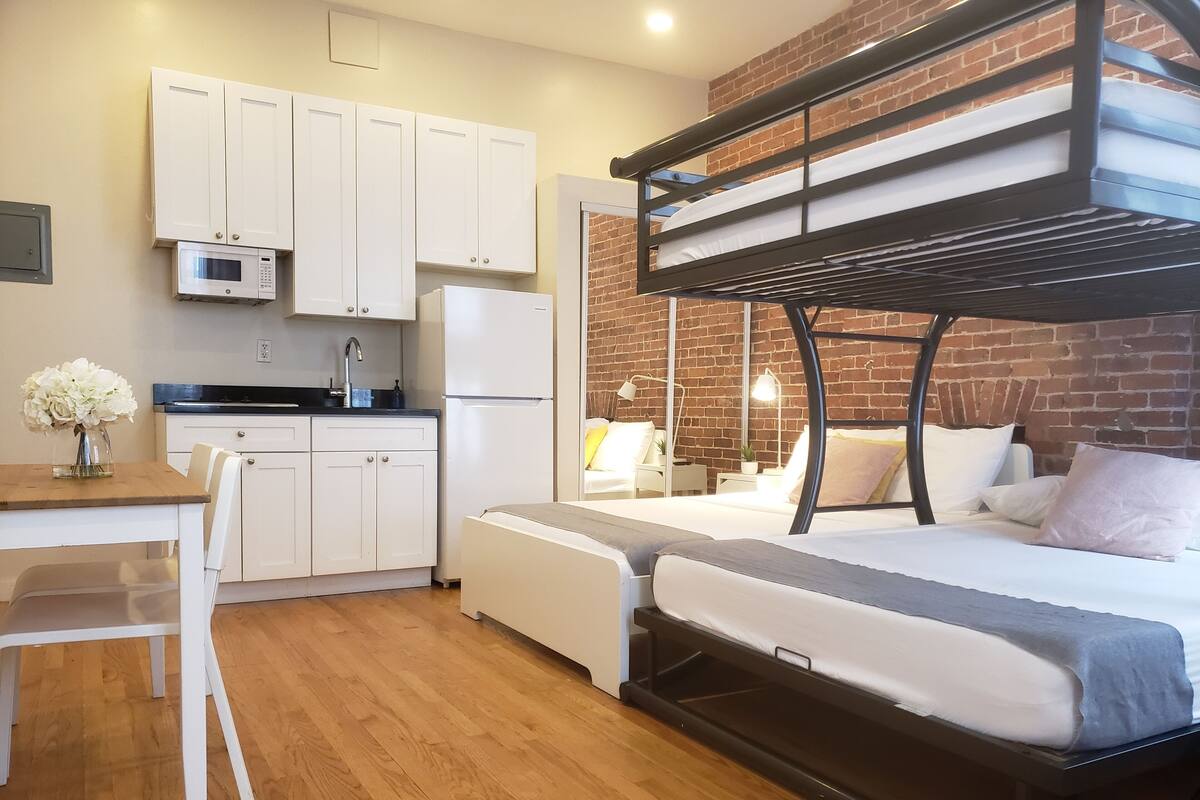 A compact studio apartment with a kitchenette, a bunk bed, a dining area, and a brick accent wall.