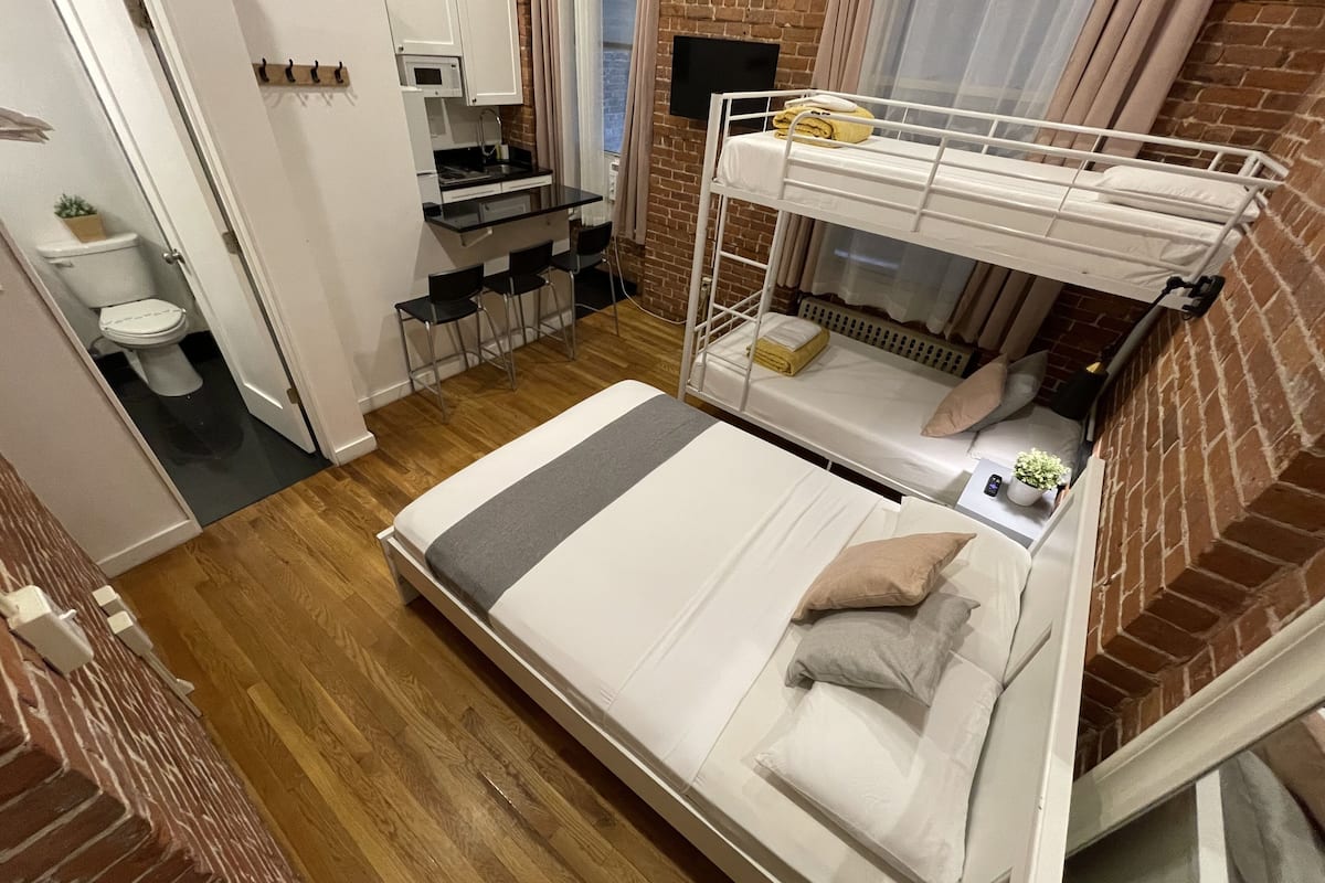 A compact hotel room with a bunk bed, a small kitchenette, a bathroom, and a view of the city.