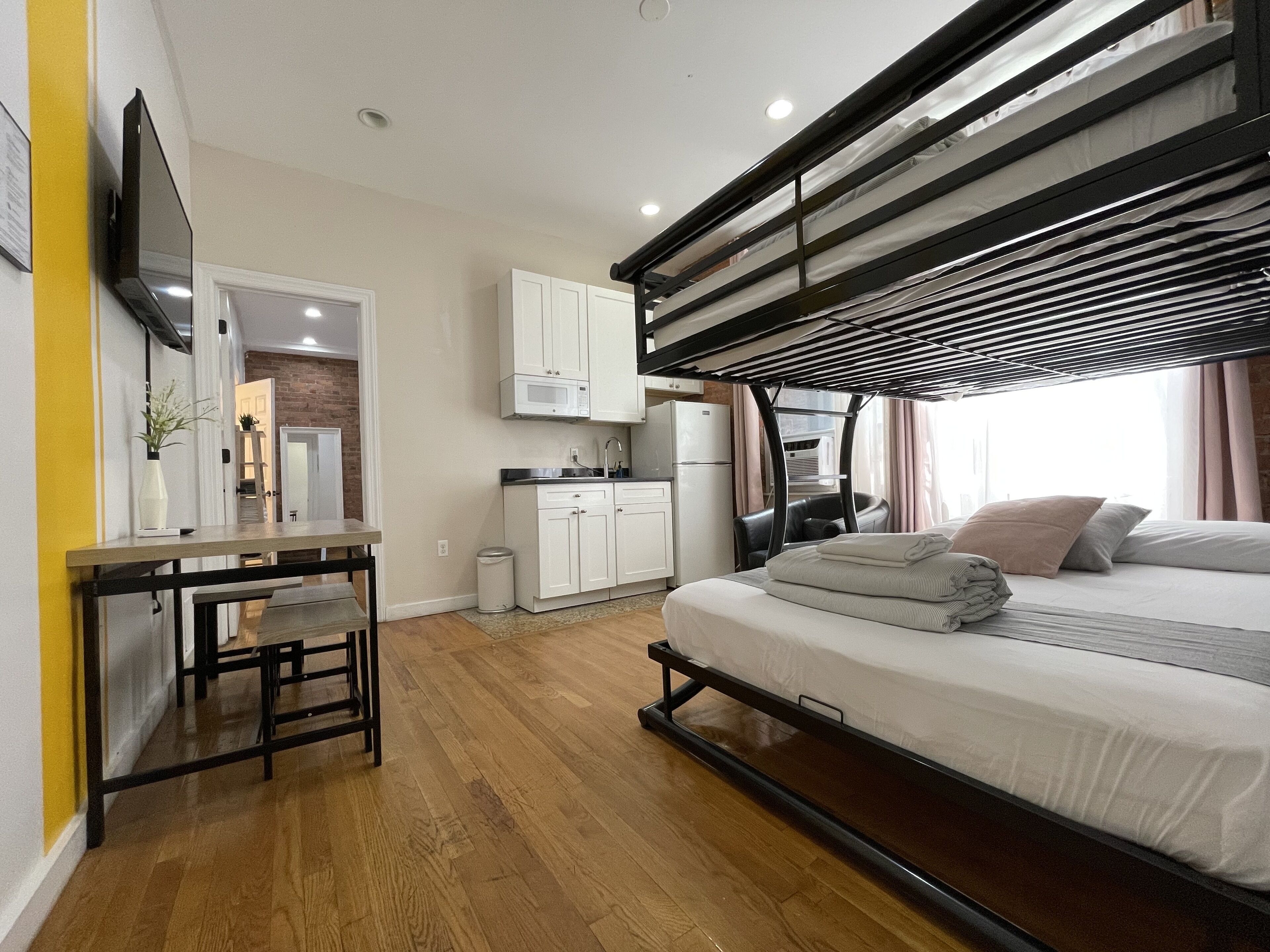 A compact studio apartment with a kitchenette, a bed with a mattress on the floor, a small table with a chair, and a TV mounted on the wall.