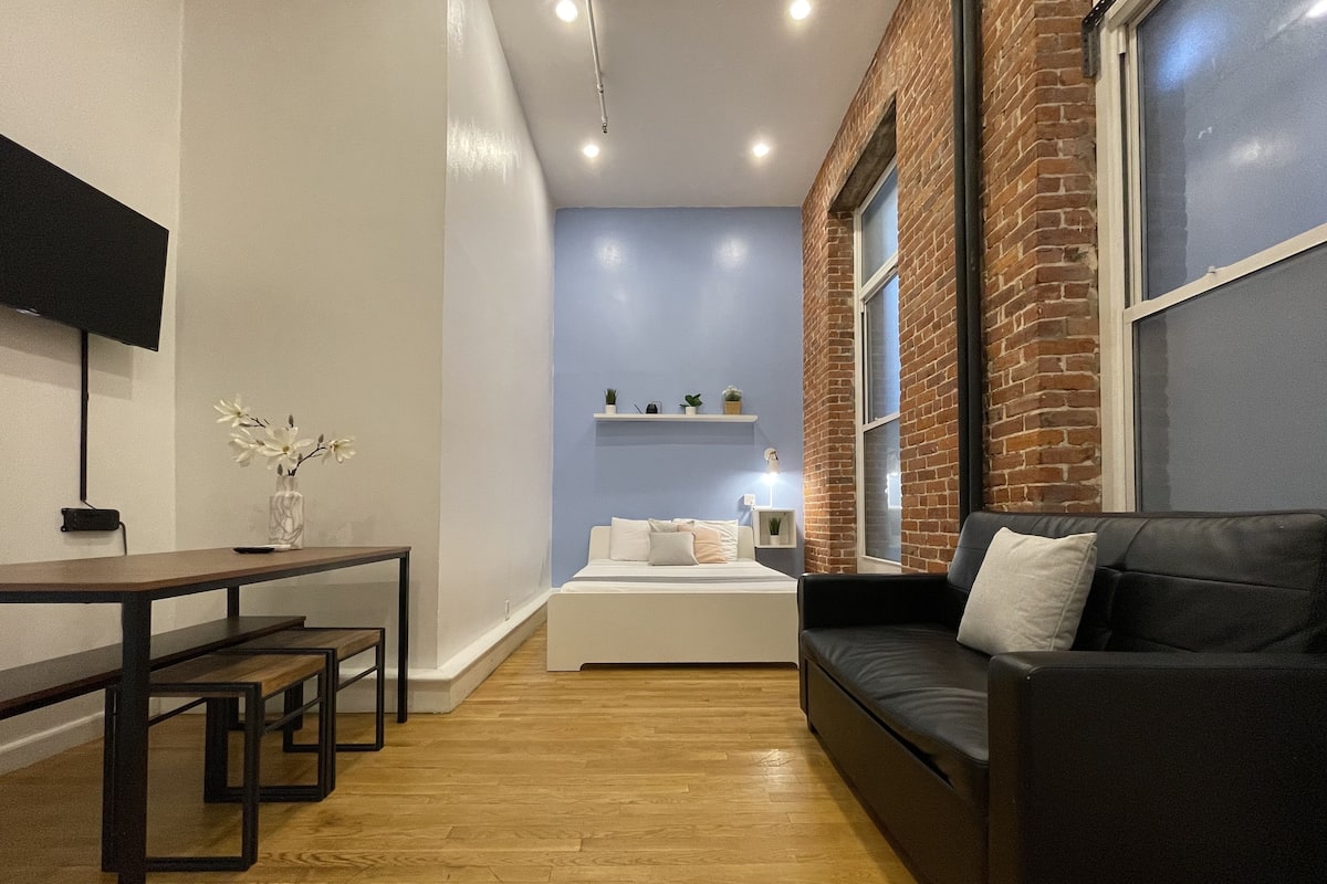 A modern living room with a black leather sofa, a wooden dining table with benches, a small white table with a vase of flowers, a bed with white bedding, and a brick wall with a window.