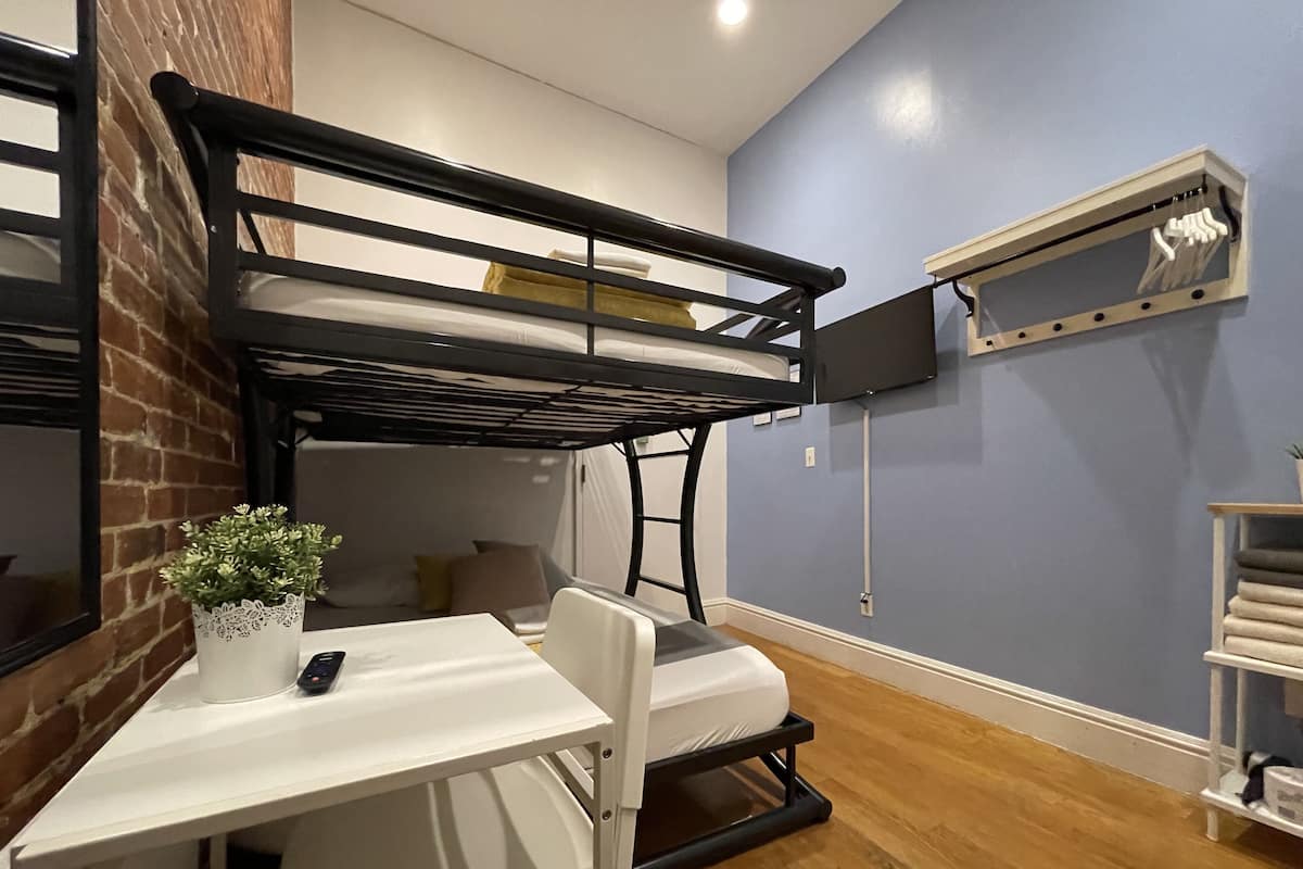 A compact room with a bunk bed, a small table with a plant, a chair, and a shelf with folded blankets.