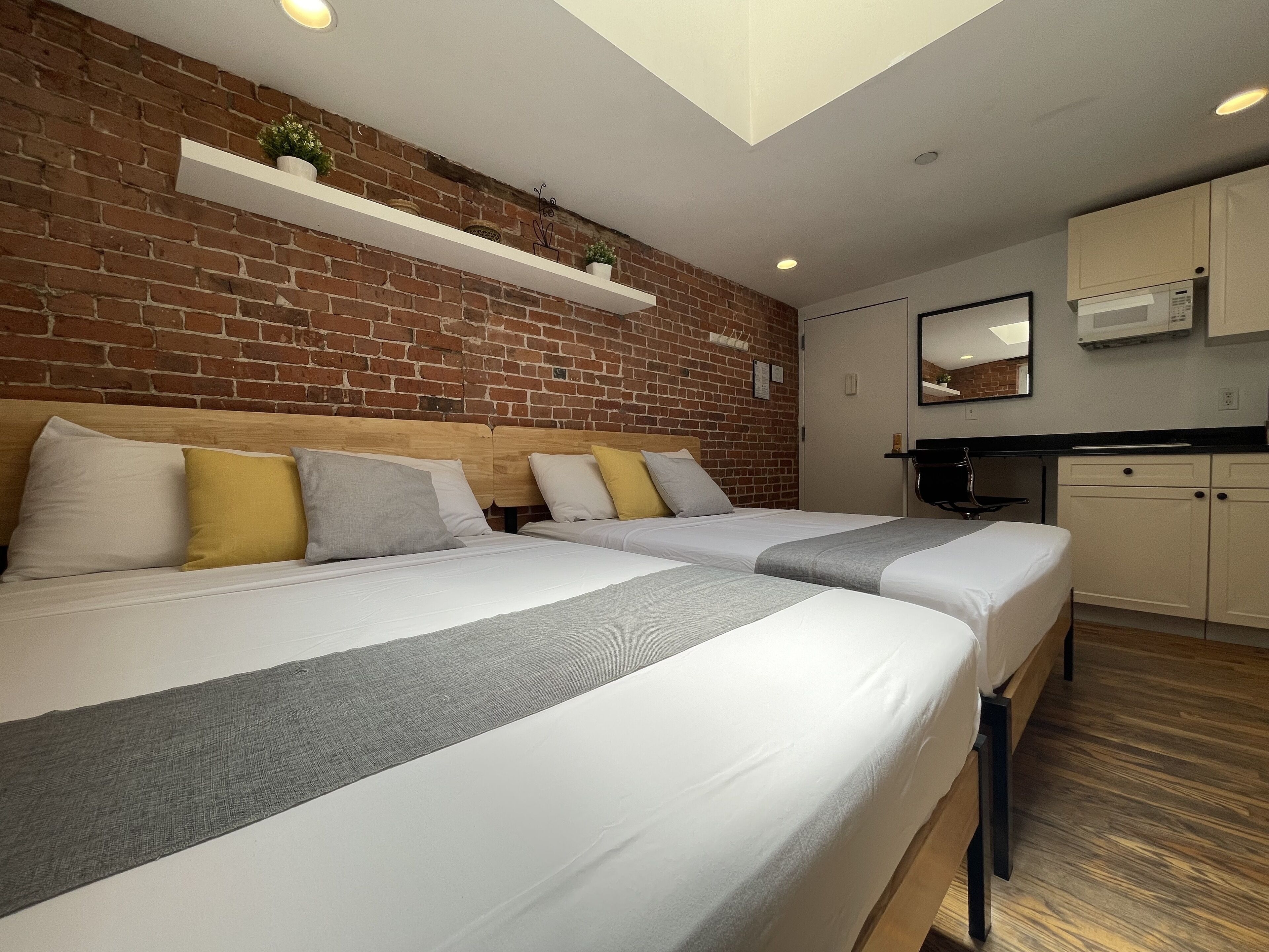Habitación de hotel con dos camas, una pared de ladrillo, una pequeña cocina y una lámpara en el techo.
