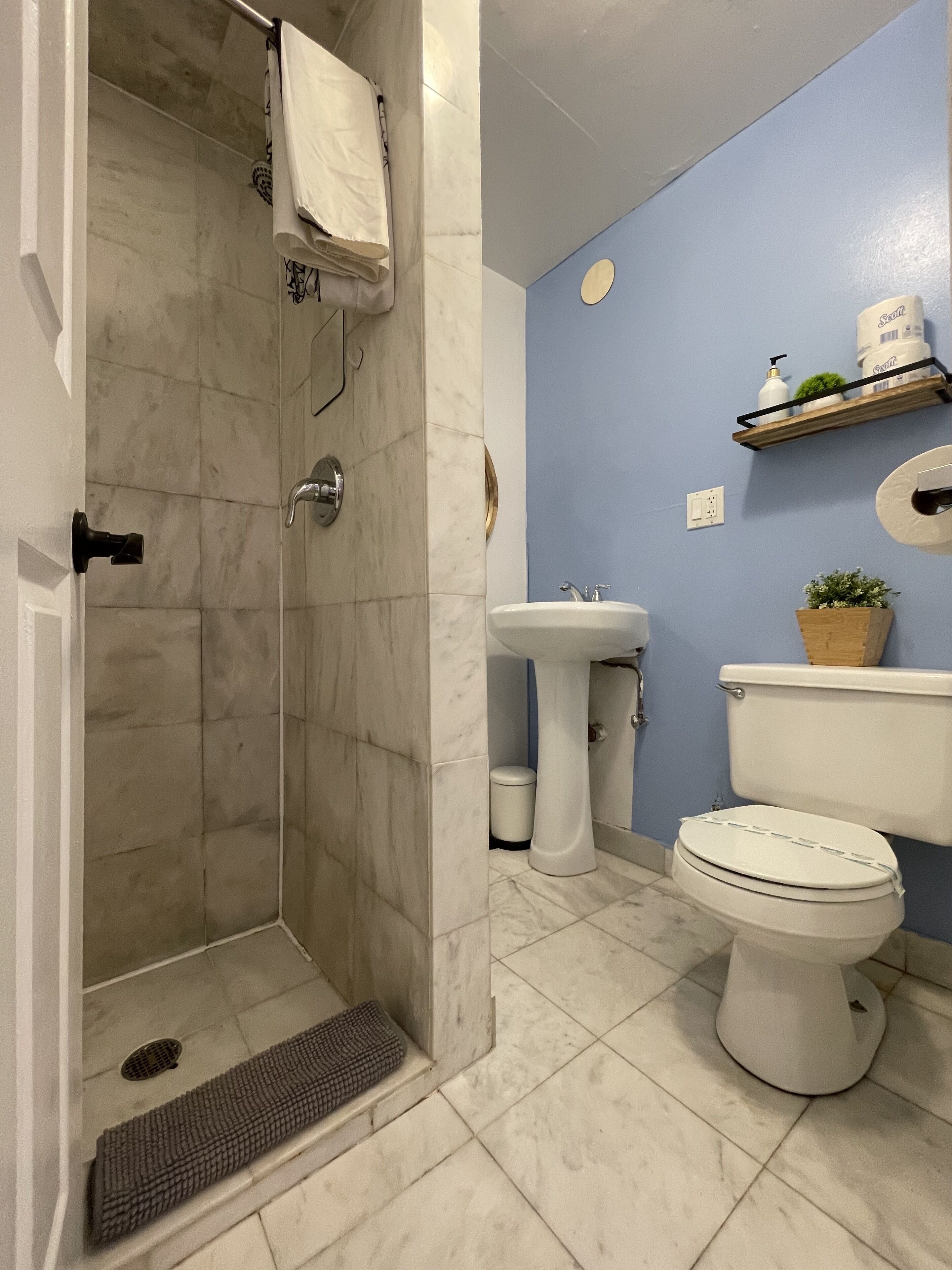 Un baño con ducha, lavamanos, inodoro y un estante con artículos de aseo.