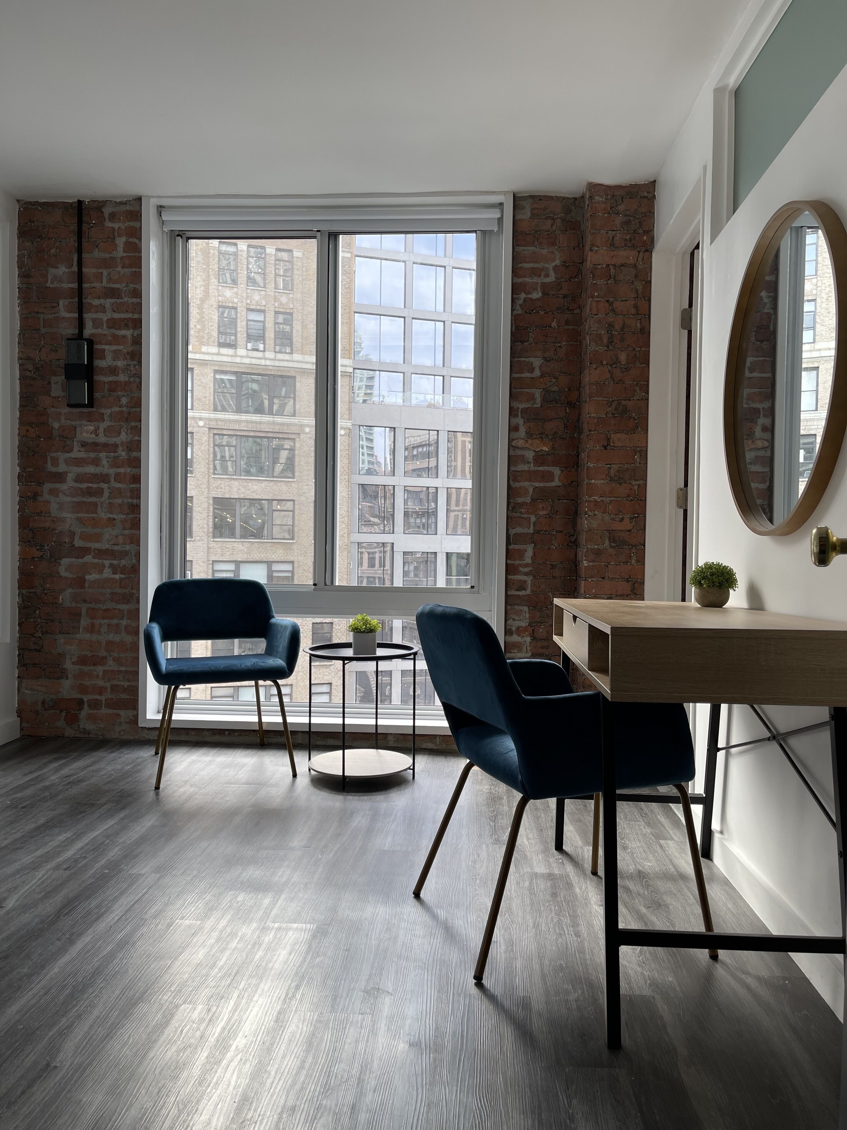 A room with a large window, two blue chairs, a small table with a plant, and a wooden desk.