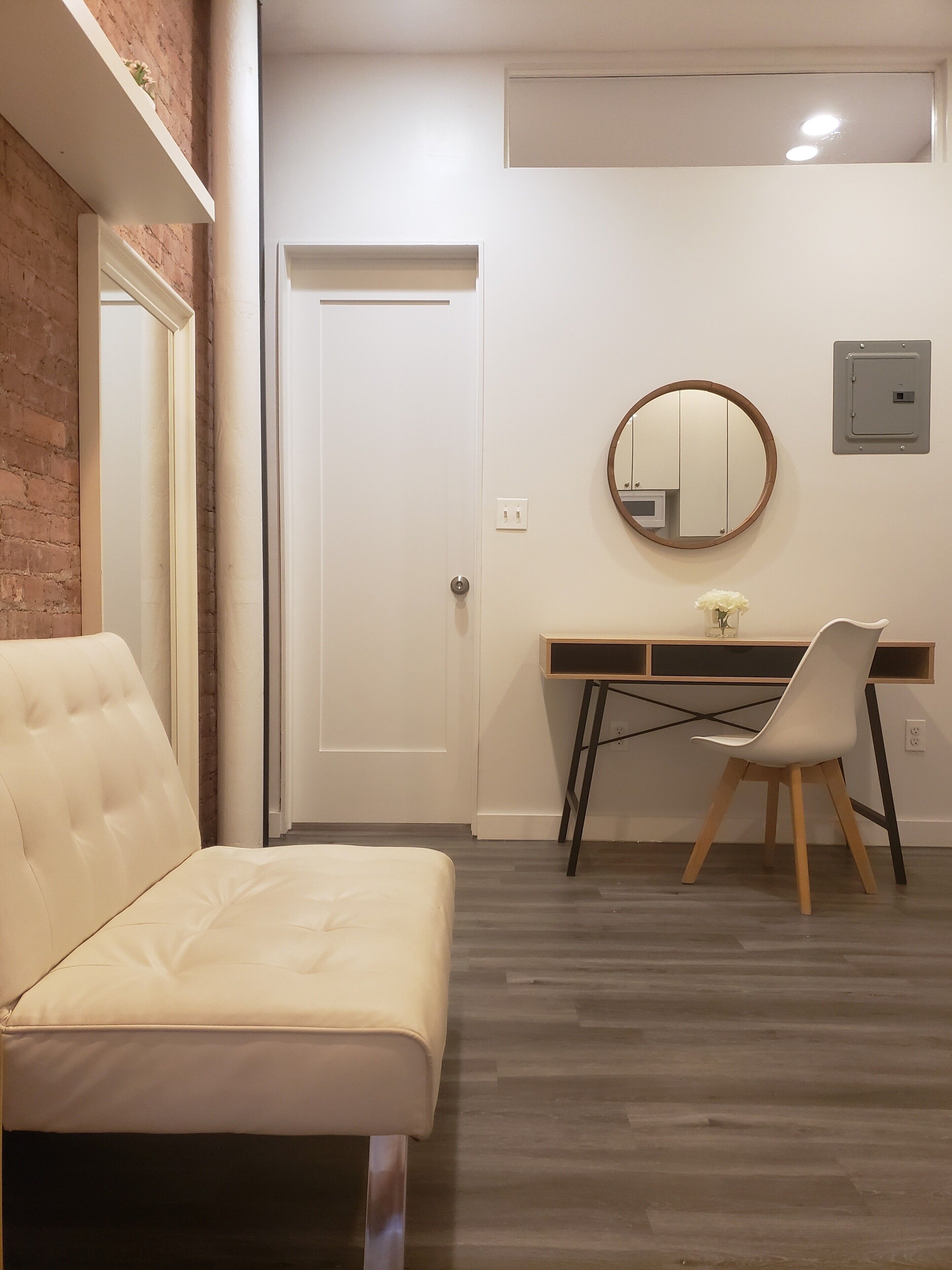 A room with a white sofa, a desk with a chair, a door, and a wall-mounted mirror.