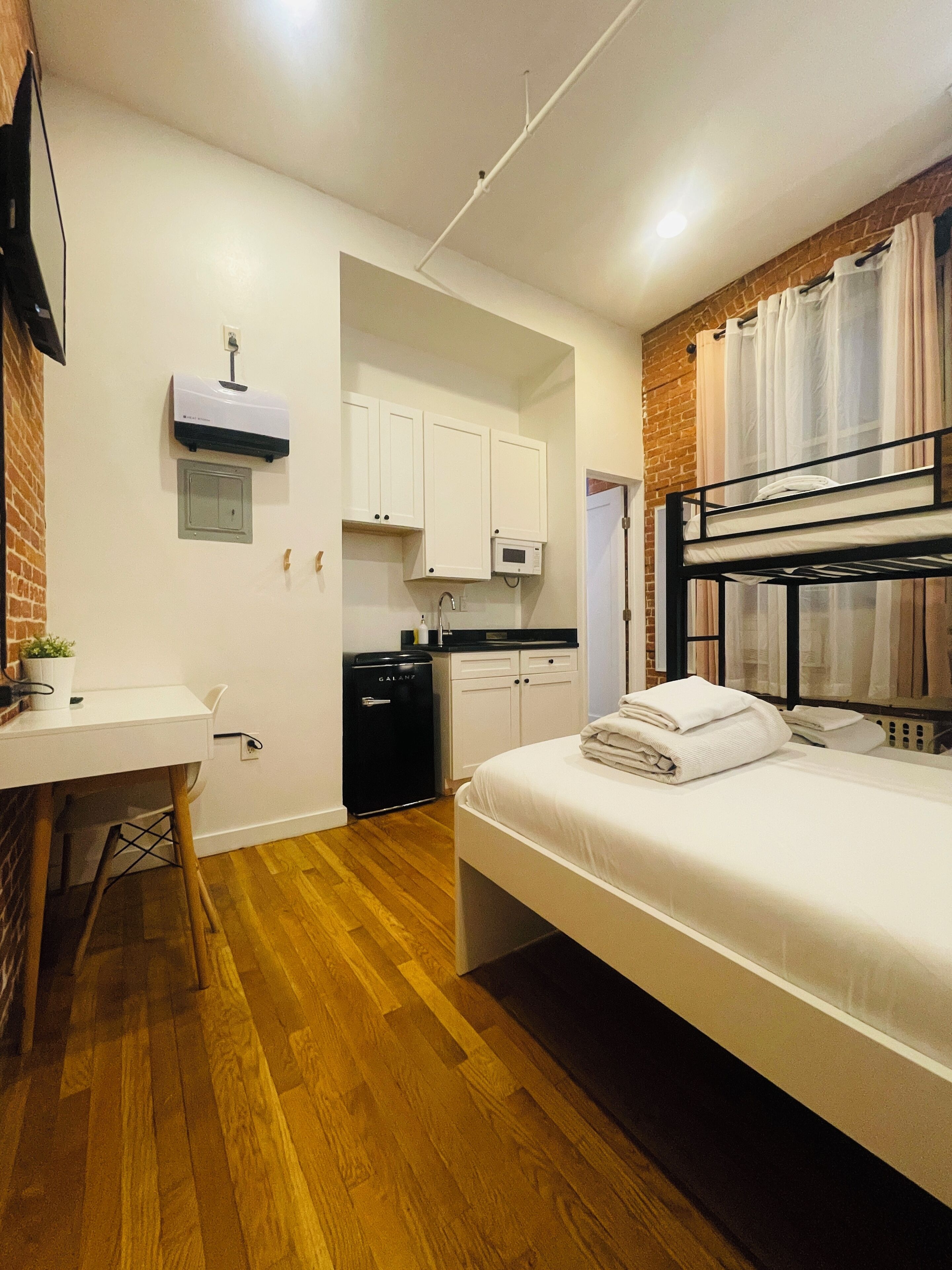 A compact hotel room with a bunk bed, a small kitchenette, a desk, and a white wardrobe.