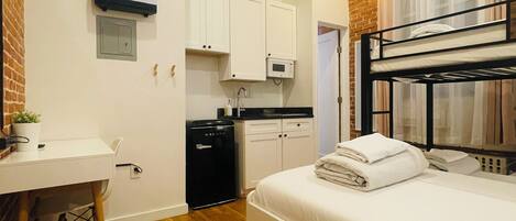 A compact hotel room with a bunk bed, a small kitchenette, a desk, and a white wardrobe.