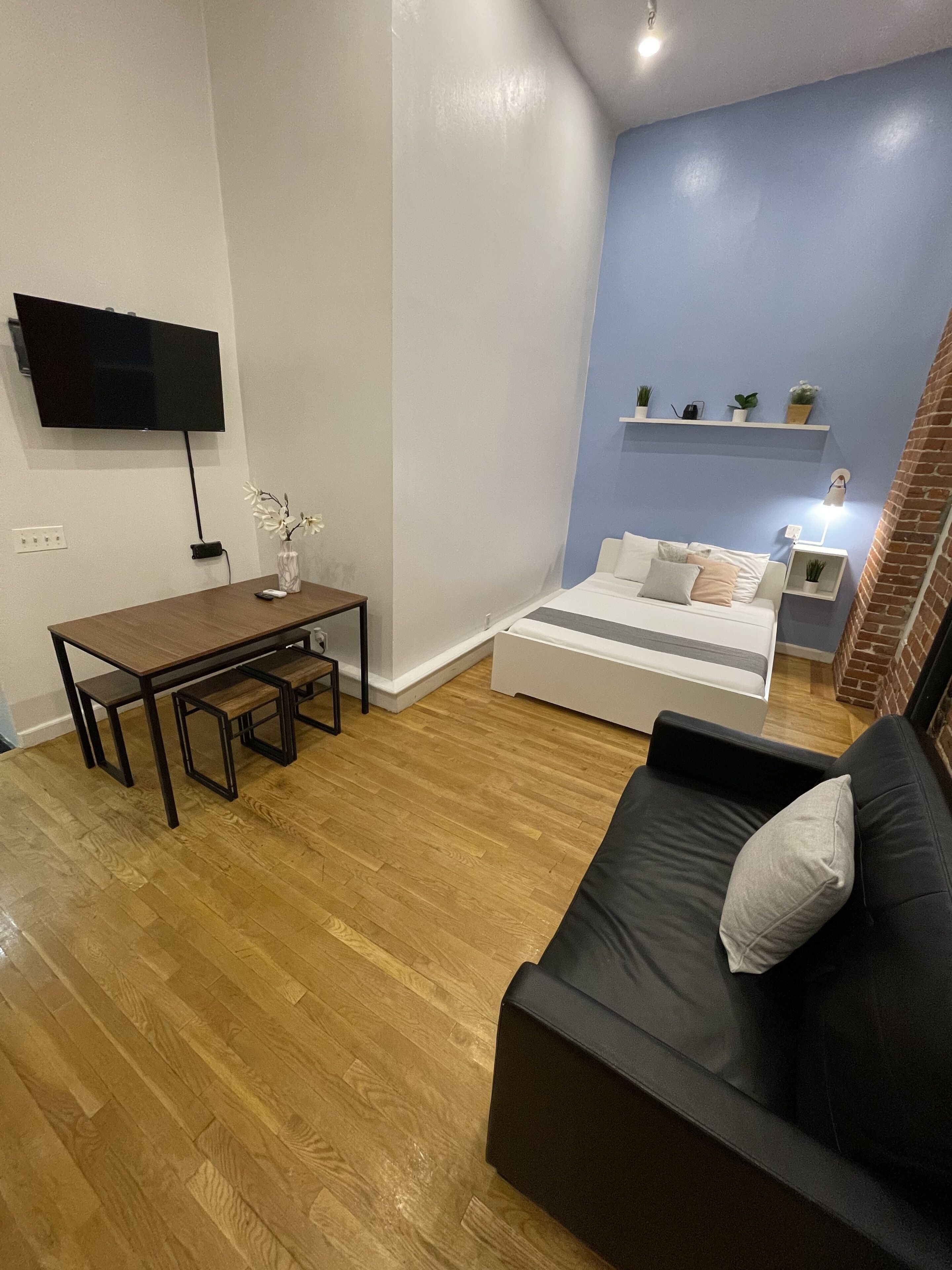 Un salón moderno con suelo de madera, un sofá negro, una cama blanca, un televisor montado en la pared y una mesita con un jarrón de flores.