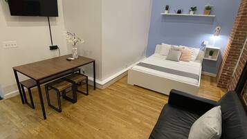 Un salón moderno con suelo de madera, un sofá negro, una cama blanca, un televisor montado en la pared y una mesita con un jarrón de flores.