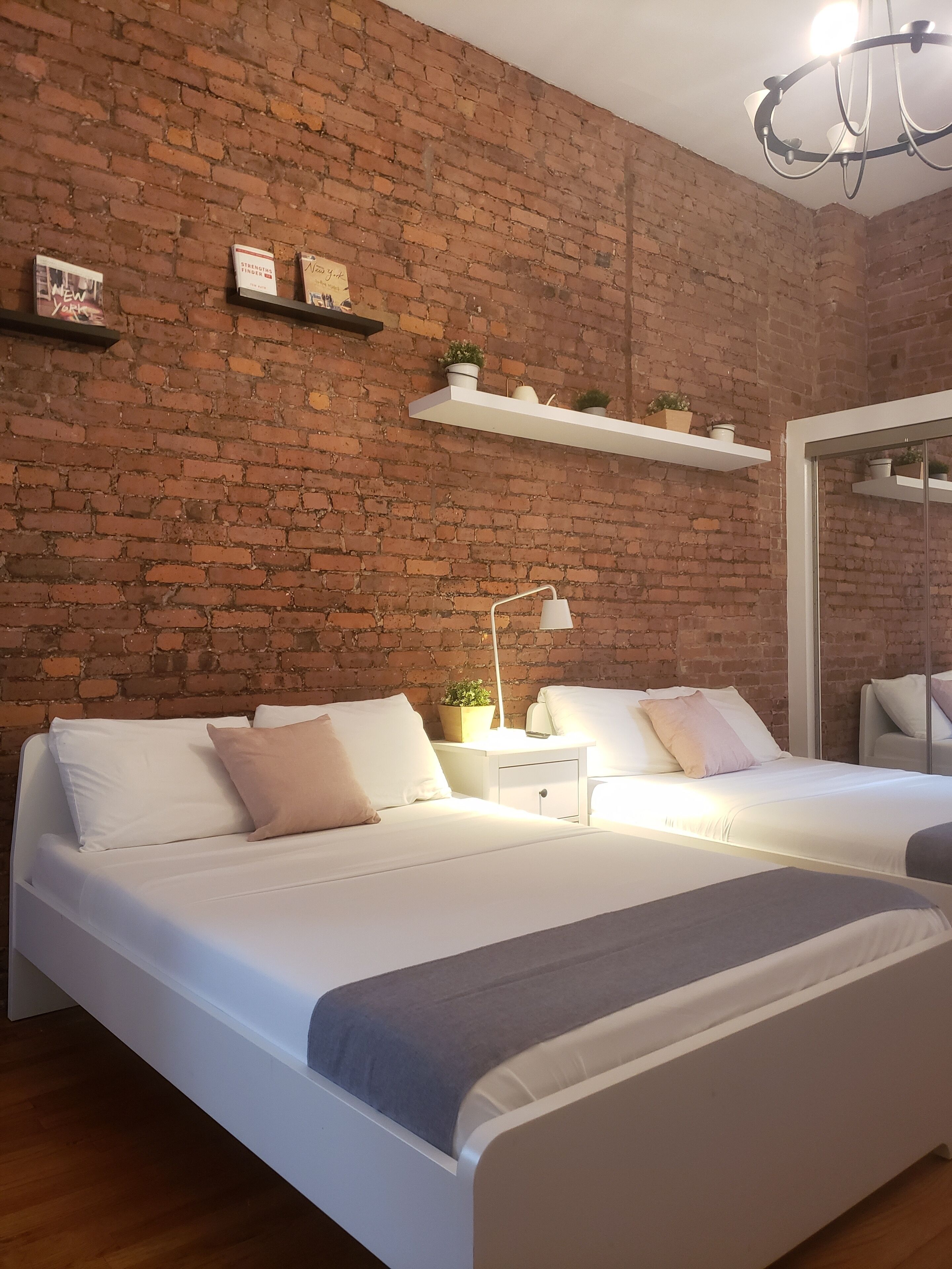 Een moderne slaapkamer met een bakstenen muur, twee bedden, wit beddengoed, een nachtkastje met een lamp en een hangende lamp.