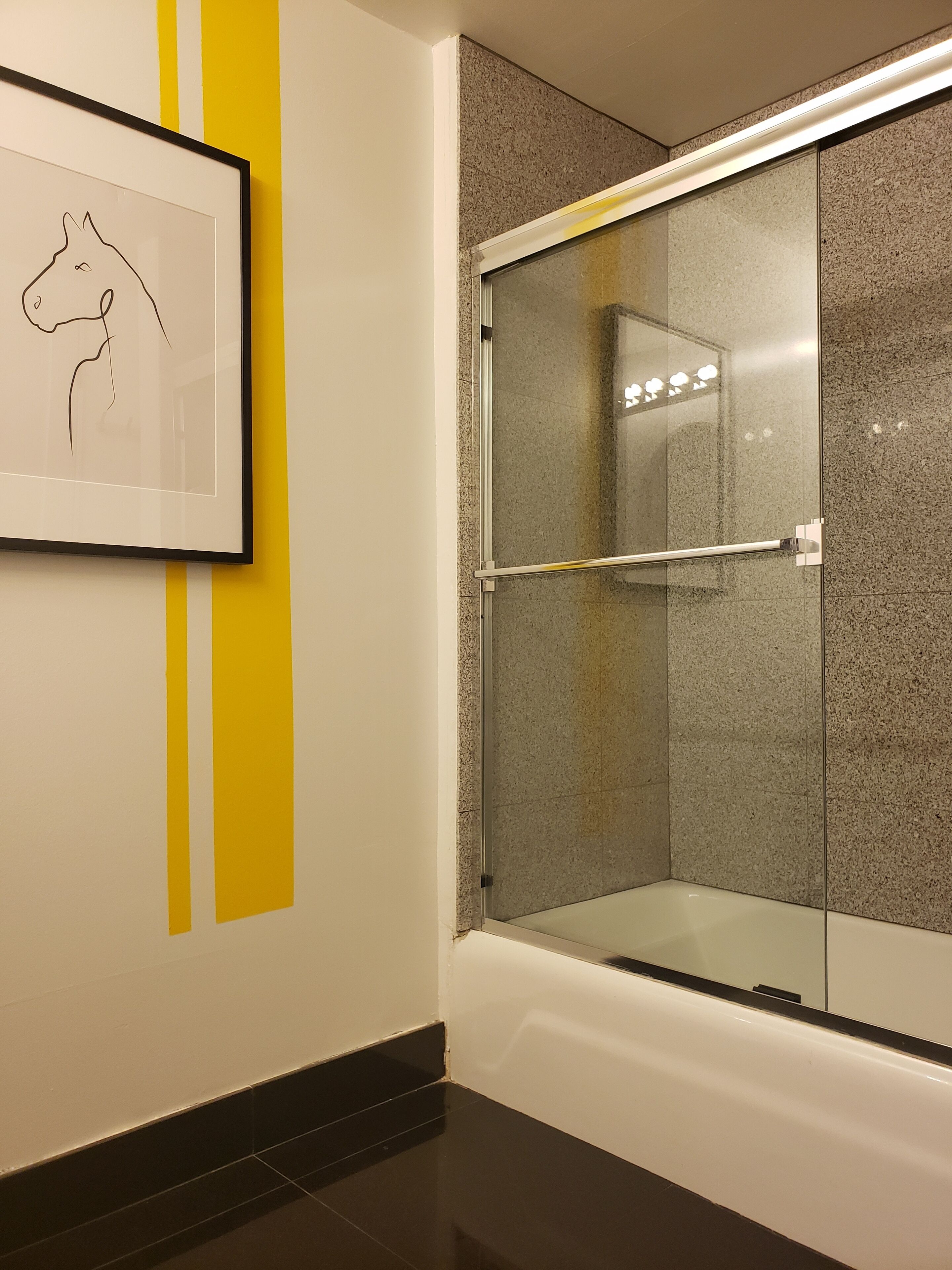 Une salle de bain moderne avec une douche à parois de verre, un dessin linéaire encadré au mur et un motif à rayures jaunes.