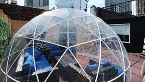 Een transparante koepelstructuur met een geometrisch frame, tegen een stadsgezicht met hoge gebouwen en een heldere lucht.
