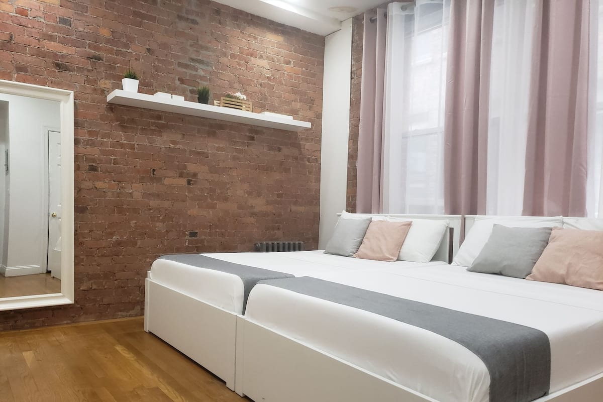 A bedroom with a brick wall, a white bed with grey and beige pillows, a white shelf with decorative items, and a window with sheer curtains.