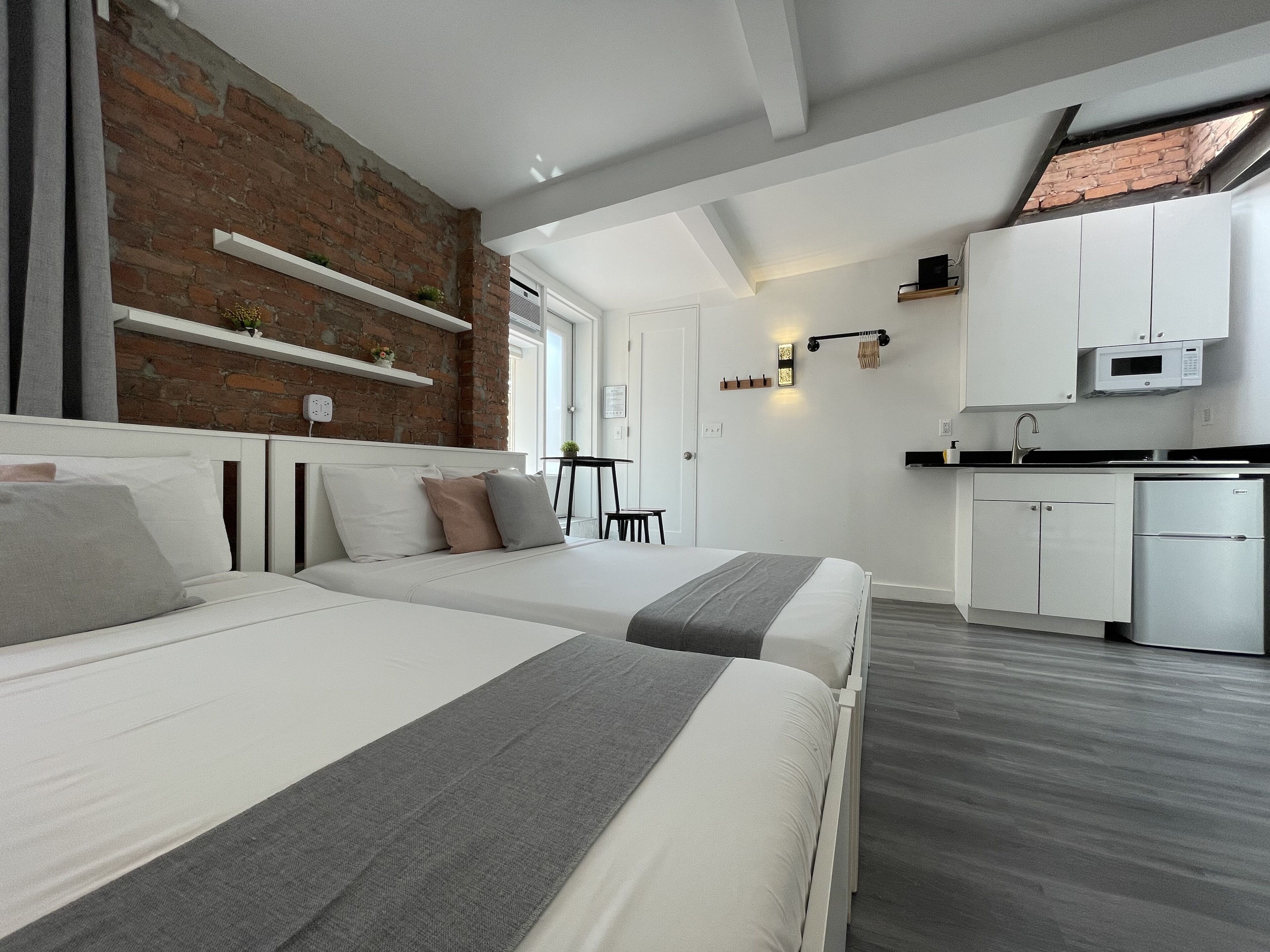 A modern hotel room with two beds, a kitchen area, and a brick wall.