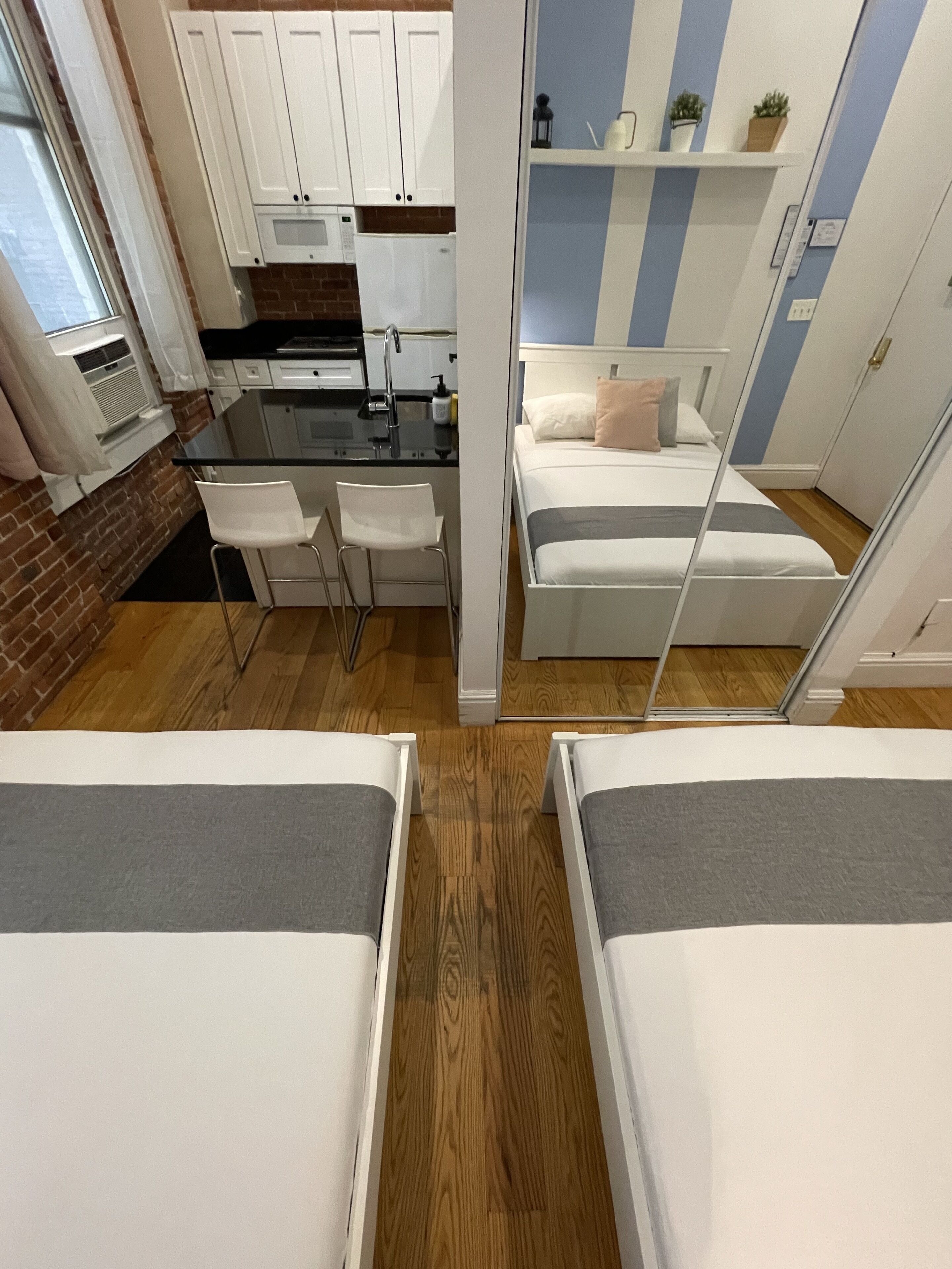 Une chambre d’hôtel compacte comprenant un coin cuisine, un petit espace repas et un lit avec une tête de lit grise.