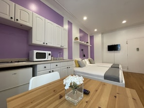 Une chambre d’hôtel compacte comprenant un coin cuisine, un lit avec des oreillers, un téléviseur fixé au mur et une petite table agrémentée d’un vase de fleurs.