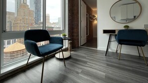 Une chambre moderne dotée d’une grande fenêtre donnant sur la ville, de deux fauteuils bleus, d’un miroir rond et d’une petite table sur laquelle est posée une plante.