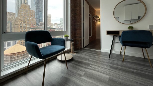 Een moderne kamer met een groot raam met uitzicht op de stad, twee blauwe stoelen, een ronde spiegel en een tafeltje met een plant.