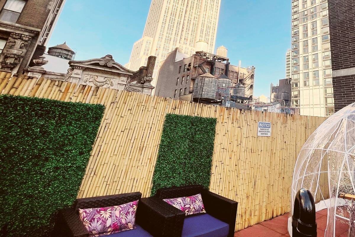 A rooftop terrace with a view of a tall building, a bamboo wall, and a seating area with cushions.