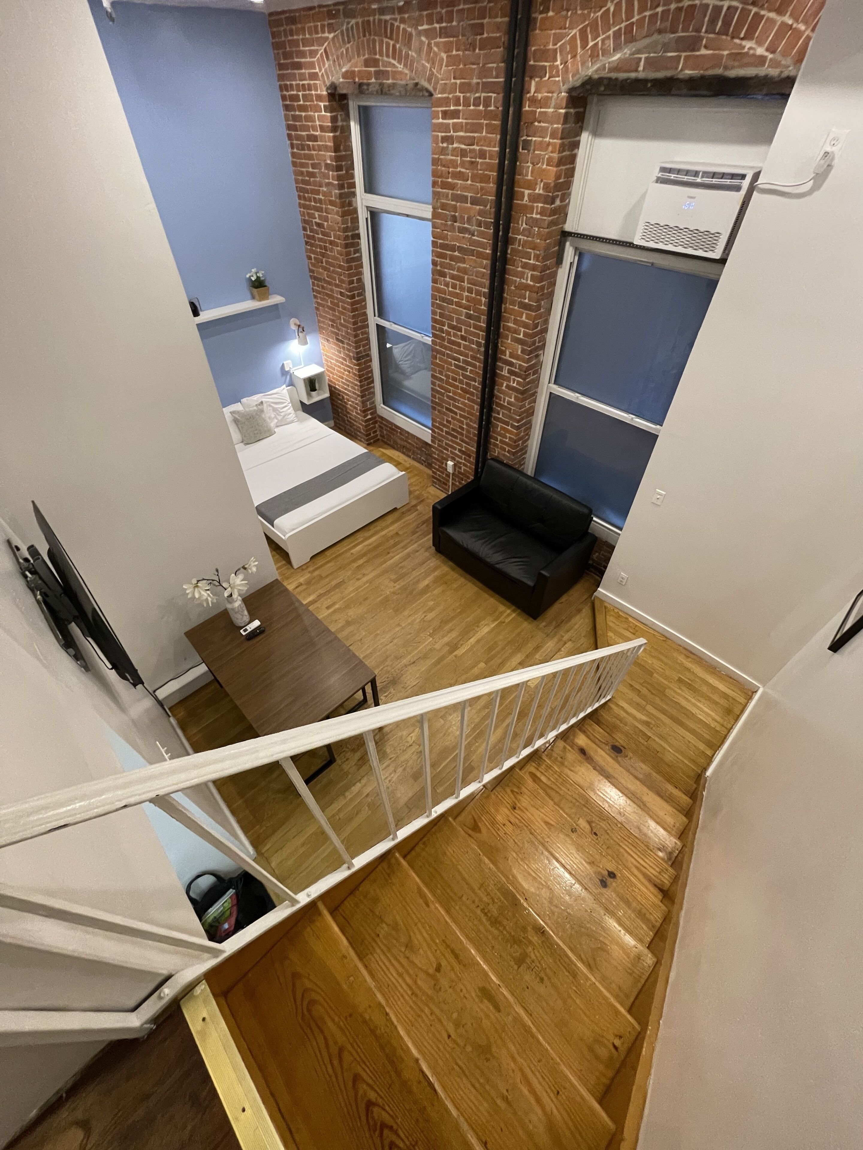 A loft-style bedroom with a brick wall, a bed, a black sofa, and a wooden staircase.