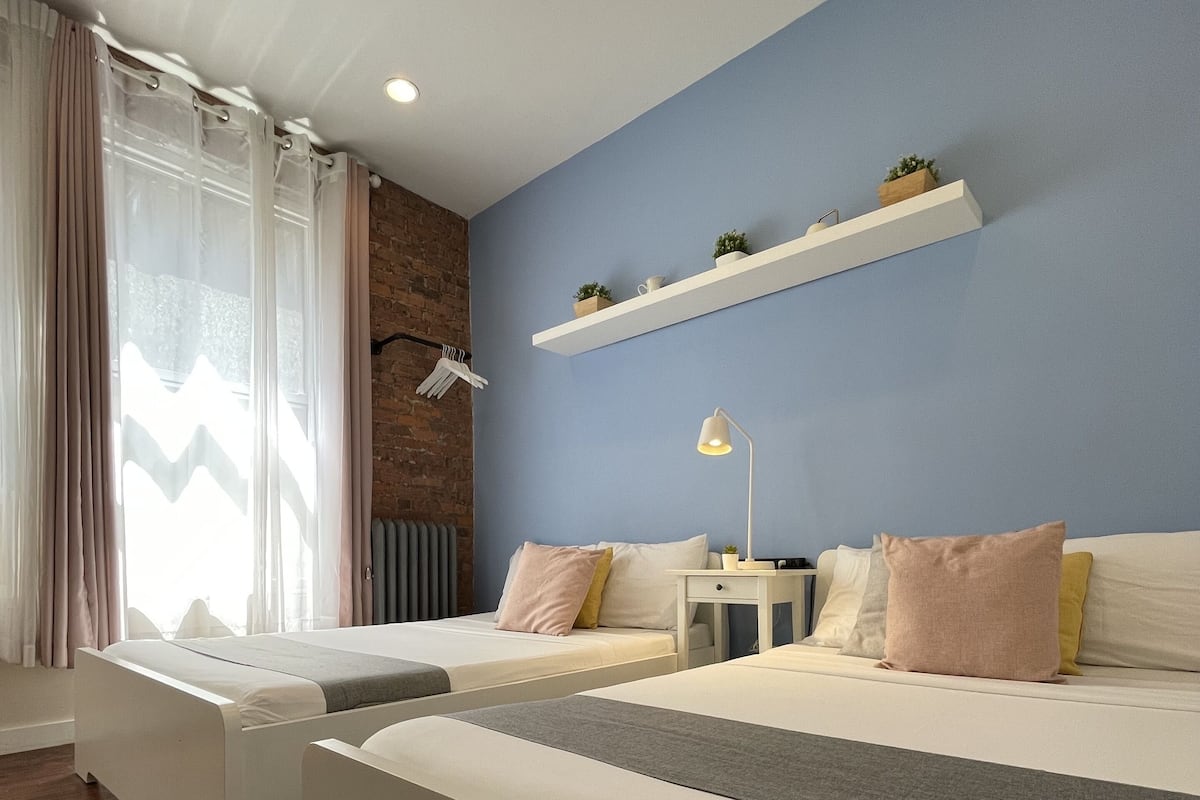 A modern bedroom with two beds, a white bench, a radiator, a wall-mounted shelf with plants, and a window with curtains.