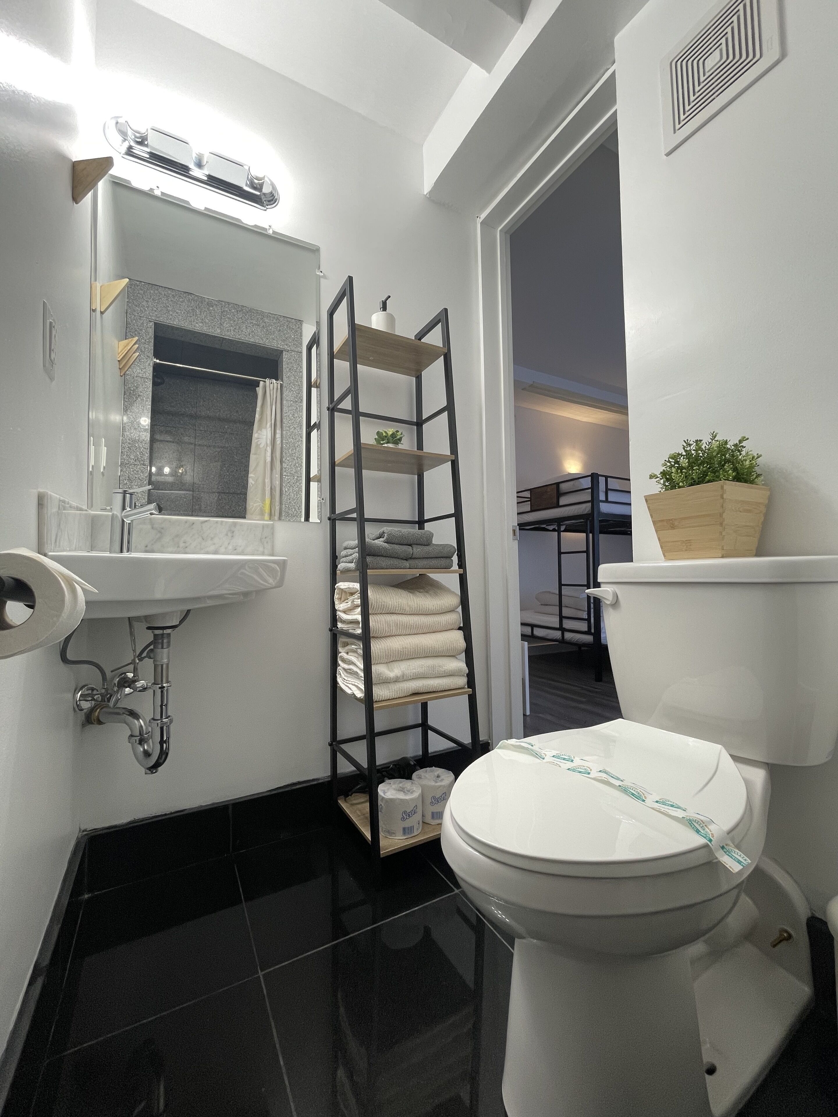 A modern bathroom with a white toilet, a black tiled floor, a wooden shelf with towels, and a mirror above the sink.