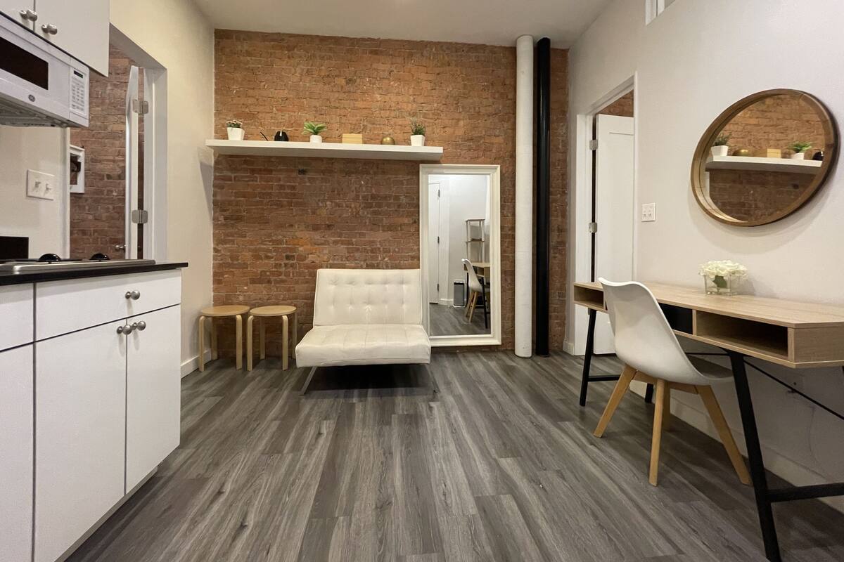 A compact room with a kitchenette, a white sofa, a desk, and a mirror.