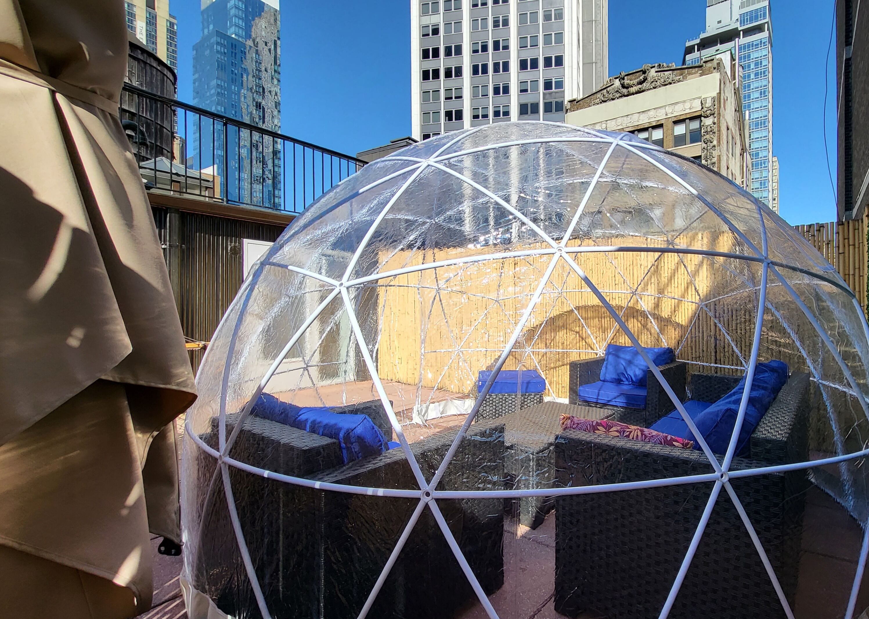Una estructura de cúpula transparente con mobiliario de mimbre en su interior, con un paisaje urbano de fondo.