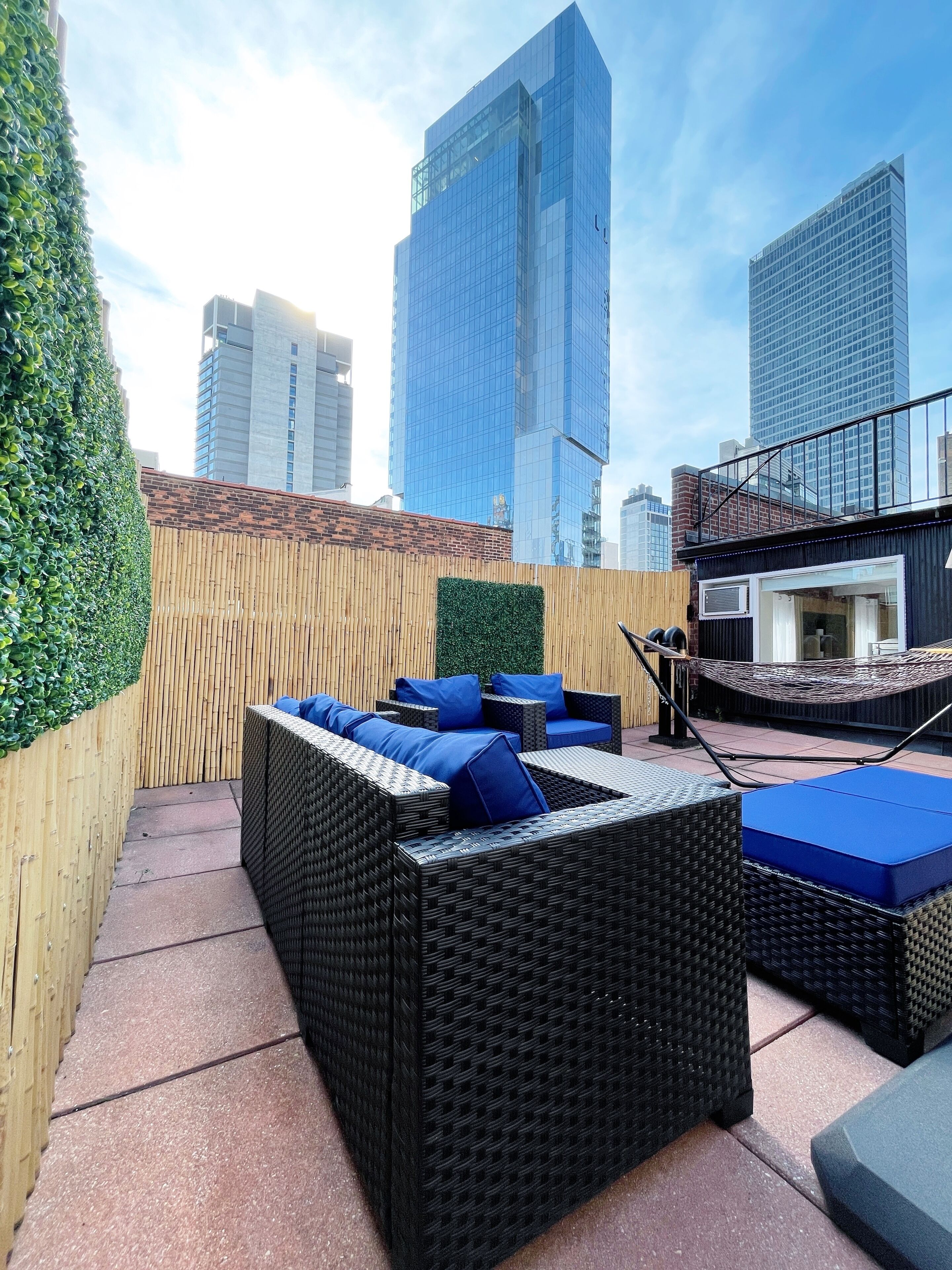 Een terras op het dak met rieten meubels en blauwe kussens, met uitzicht op een stadsgezicht met hoge gebouwen.
